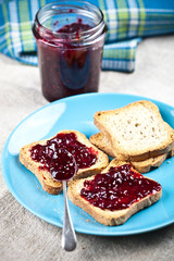 Toasted cereal bread slices on blue ceramic plate and homemade wild berries jam in jar, spoon and linen napkin closeup on rustic wooden table background.