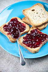 Fresh toasted cereal bread slices with homemade wild berries jam and spoon on blue ceramic plate closeup on linen tablecloth background.