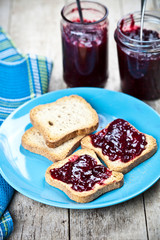 Fresh cereal bread slices on blue ceramic plate, homemade cherry and wild berries jam in jars and spoons closeup on linen napkin on rustic wooden table background.
