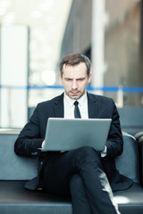 Serious middle aged businessman in suit sitting on leather couch in airport lounge or office lobby and working on laptop computer placed on his lap