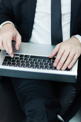 Close-up high angle view of hands of anonymous businessman in suit typing on laptop computer placed on his lap sitting on leather sofa
