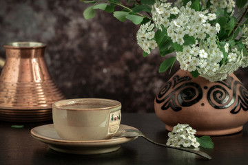 On the table is a cup of coffee and a vase with white flowers of spirea