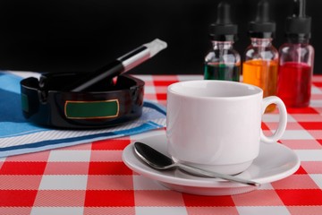 cup of coffee and electronic cigarette in ashtray with bottles of vape liquid on checkered tablecloth