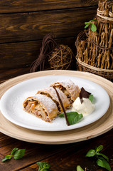 Apple strudel with creamy ice cream, garnished with mint on a white plate on a short wooden background. Close-up. Space