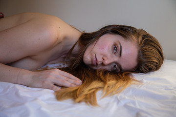 Naklejka premium Portrait of young lonely naked pretty woman with brown hair lying on bed and looking at camera