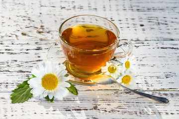 A cup of tea and a saucer, standing on a wooden, white table, outdoors, in the rays of sunlight and the flowers of chamomile white.