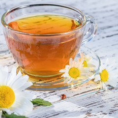 A cup of fragrant tea standing on a wooden, white table, in the rays of sunlight, in the flowers of the field chamomile.