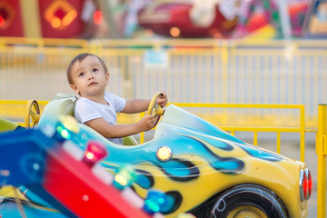 Little toddler in theme park: a boy in white shirt riding little car on merry go round, bright...