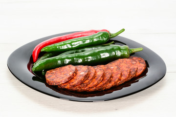 Pepper salami in slices with some red and green chilli fresh peppers on glossy black plate on a white wooden table.