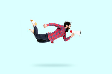 Bearded hipster man in glasses, jumping and shouting in megaphone, photo over blue background
