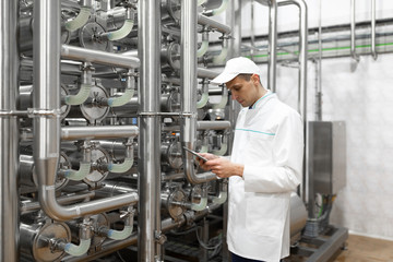 Portrait of man in a white robe and a cap standing in production department of dairy factory with grey tablet