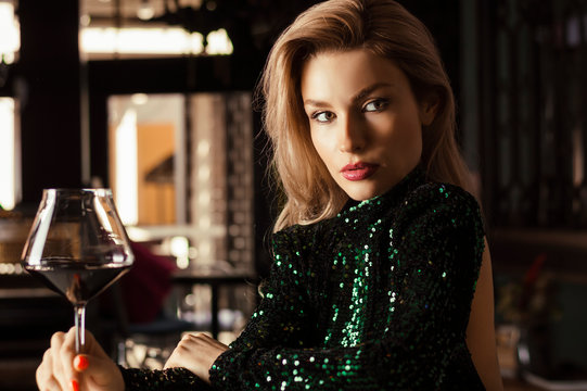 Seductive blonde in a green evening dress posing with a glass of red wine.