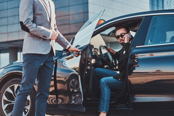Busy nice businessman in sunglasses is talking by smartphone while his elegant assistant is opening...