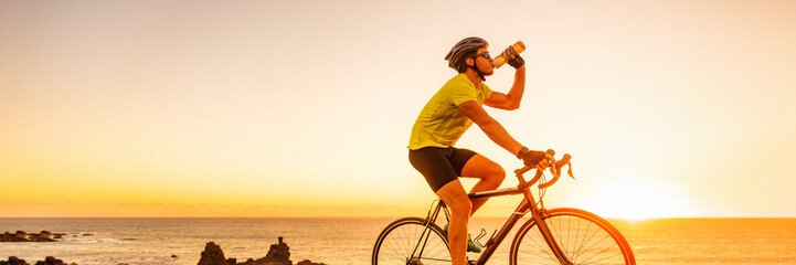Triahtlon athlete man drinking water bottle on road racing bike ride outdoors at sunset banner panorama landscape. Cyclist biking outside with sunglasses and helmet.