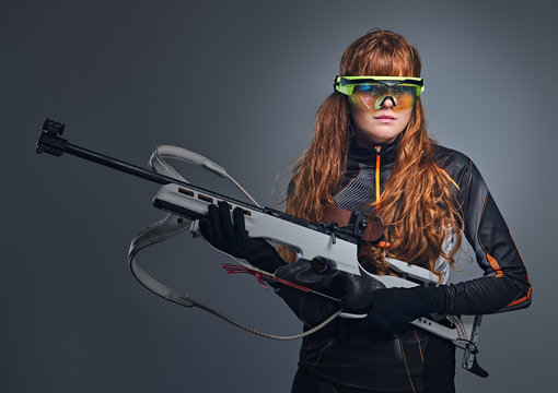 Redhead Biatlon female sportsmen in eyeglasses holds competitive gun.