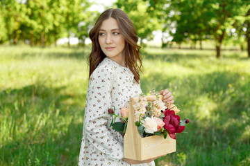 Naklejka na ściany i meble Half portrait of a charming positive woman dressed in long white summer dresses with a happy smile on the background of a green meadow in the hands of a wooden basket with peonies, roses, green lilies
