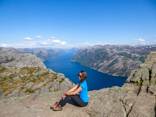 Naklejka premium A girl wearing blue T-shirt sitting at the edge of a steep cliff of Preikestolen, with a view on Lysefjorden. Fjord goes far inland. Girl enjoys the view, feeling free and happy. Great accomplishment