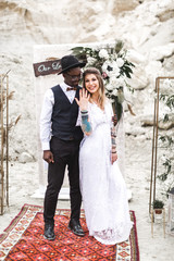 African groom and Caucasian bride embracing each other in a boho style before the wedding arch from...