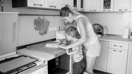 Black and white image of young mother teaching her 3 years old toddler son cooking and baking cookies on kitchen