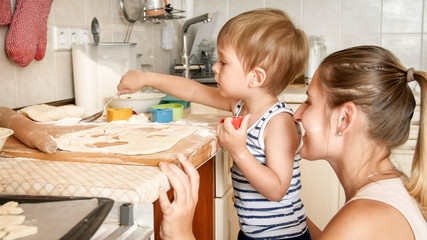 Closeup portrait of adorable 3 years old toddler boy making cookies with mother. CHild holding baking pan and putting cookies on it