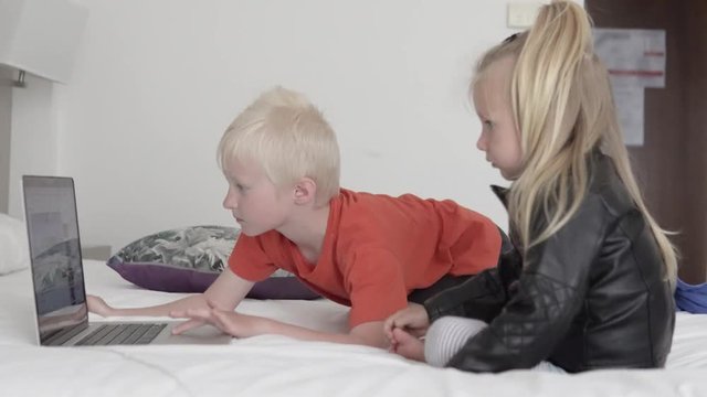 Children watch a cartoon on the computer. Brother and sister sit on the bed in the hotel and watch a cartoon