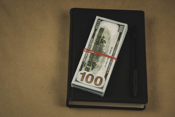 A black notebook , a pen and dollar cash banknotes on wooden background - concept of financial management or planning, make money from freelance writing