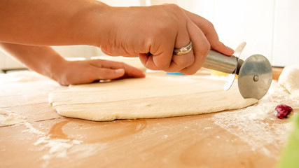Closeup image of female hand holding round knife for pizza and cutting dough on big wooden desk on...