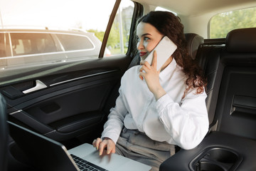 Female entrepreneur travelling to office in a luxurious car sitting on backseat with laptop and looking outside the window while talking on phone.