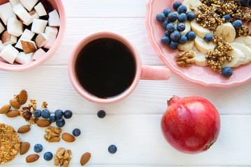 Fresh tasty fruits: coconut, bananas and blueberry and a cup of black coffee on white table. Pastel pink plates and cup, red pomegranate on white wooden table. 