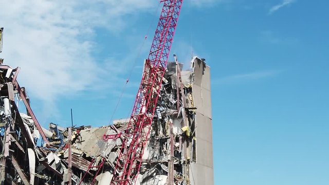 Wrecking ball on a red crane  is lowered onto a large partially demolished building. A portion of the top outside wall cracks and falls from the building. The sky is deep blue with  puffy clouds