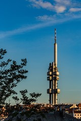 Prague, Czech Republic - May 21 2019: Aerial view of Zizkov television tower from Vitkov hill on a sunny spring evening. Blue sky with clouds. Green tree in foreground. Vertical image.