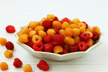 Red and yellow raspberries are located in a plate on a white background