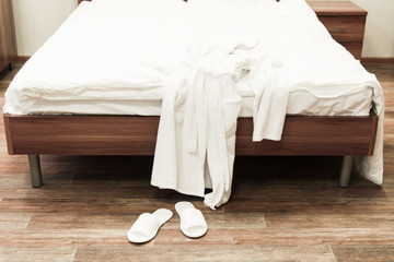 Fototapeta na wymiar Bed with white linens and white bathrobe with slippers in hotel
