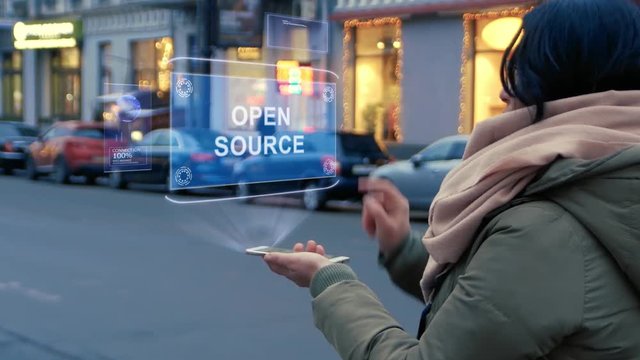 Unrecognizable woman standing on the street interacts HUD hologram with text Open source. Girl in warm clothes uses technology of the future mobile screen on background of night city