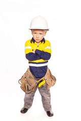 Young blond caucasian boy arms folded role playing as an angry construction worker in a yellow and blue hi-viz shirt, boots, white hard hat, tool belt, a hammer and tape measure.