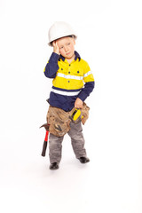 Young blond caucasian boy scratching his head in disbelief role playing a frustrated construction worker in a yellow and blue hi-viz shirt, boots, white hard hat, tool belt, a hammer and tape measure