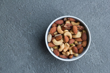 Mixed nuts (almonds, cashews, pecans, and pistachios) in a white bowl against grey cement background. Healthy snacks concept.