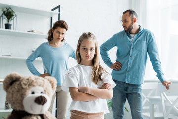 selective focus of cute kid standing with crossed arms near teddy bear and parents at home