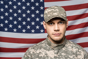 handsome soldier in military uniform and cap looking at camera near flag of america