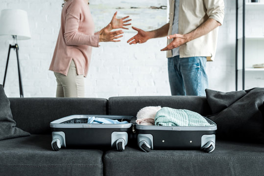 cropped view of man and woman standing and gesturing near suitcase with clothes on sofa