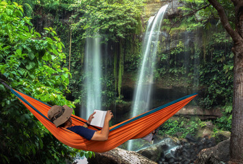 Man lying in a hammock in forest and enjoying a book reading. Man by the waterfall hanging on...
