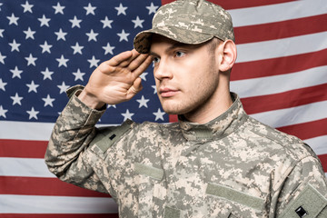 handsome soldier in military uniform and cap giving salute near flag of america