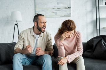 angry man sitting on sofa and screaming at upset woman - obrazy, fototapety, plakaty