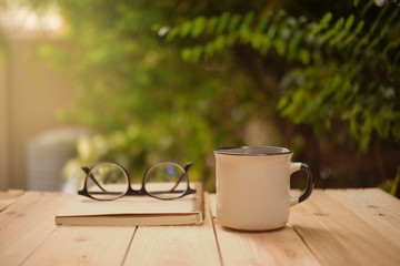 Cup of hot coffee, reading glasses and a book on wooden table in the green garden this morning. 