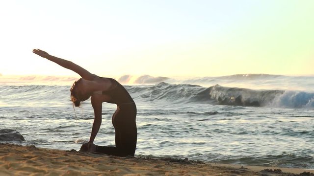 wonderful brunette in black practices yoga in core back bending pose on sand beach against rolling blue ocean waves in morning slow motion. Concept fitness sport yoga health lifestyle