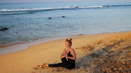 exciting lady in black sits on sand beach puts hands in Salutation Seal meditating against blue foaming ocean waves slow motion. Concept spiritual practices wellness lifestyle teacher