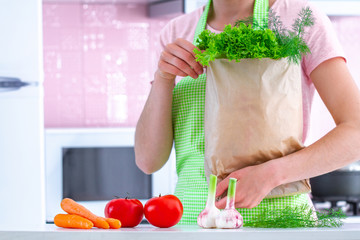 Cooking woman in apron holding a craft paper bag full of fresh organic vegetables at kitchen. Healthy food and balanced diet, clean eating