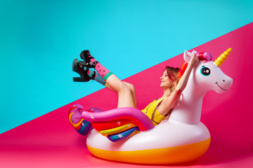 Concept summer mood, relaxation and beauty.Modern portrait of a young woman in a yellow swimsuit, bright socks and sandals resting on an inflatable unicorn mattress on an isolated pink-blue background