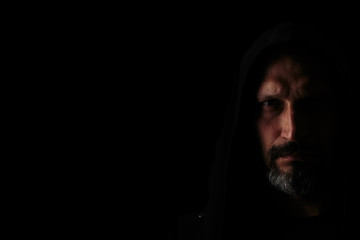 Brutal man with a gray beard in the hood with sharp shadows on a black background. Copy space. - obrazy, fototapety, plakaty