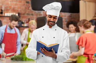 cooking class, profession and people concept - happy male indian chef in toque reading cookbook over group of students background